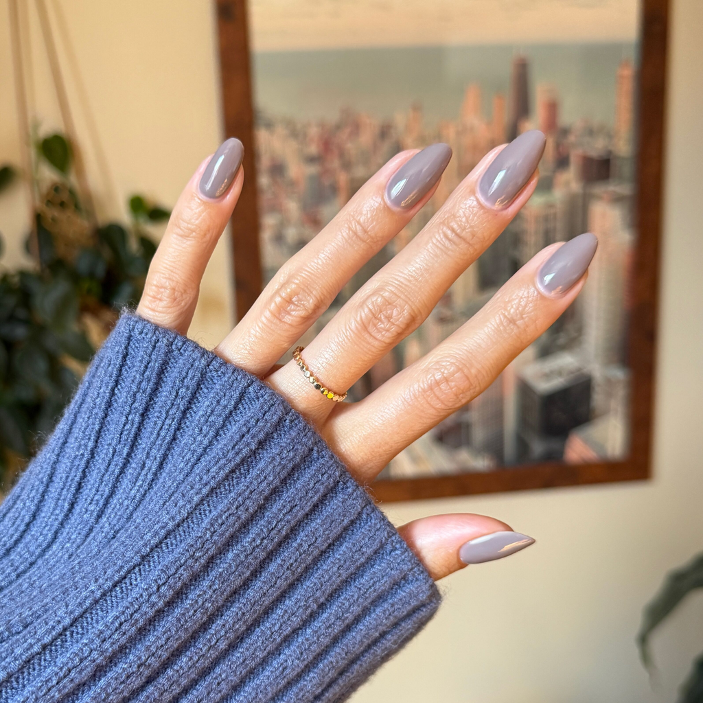 Hand with almond-shaped nails painted in glossy AW - Gel warm grey polish held against blurred city background