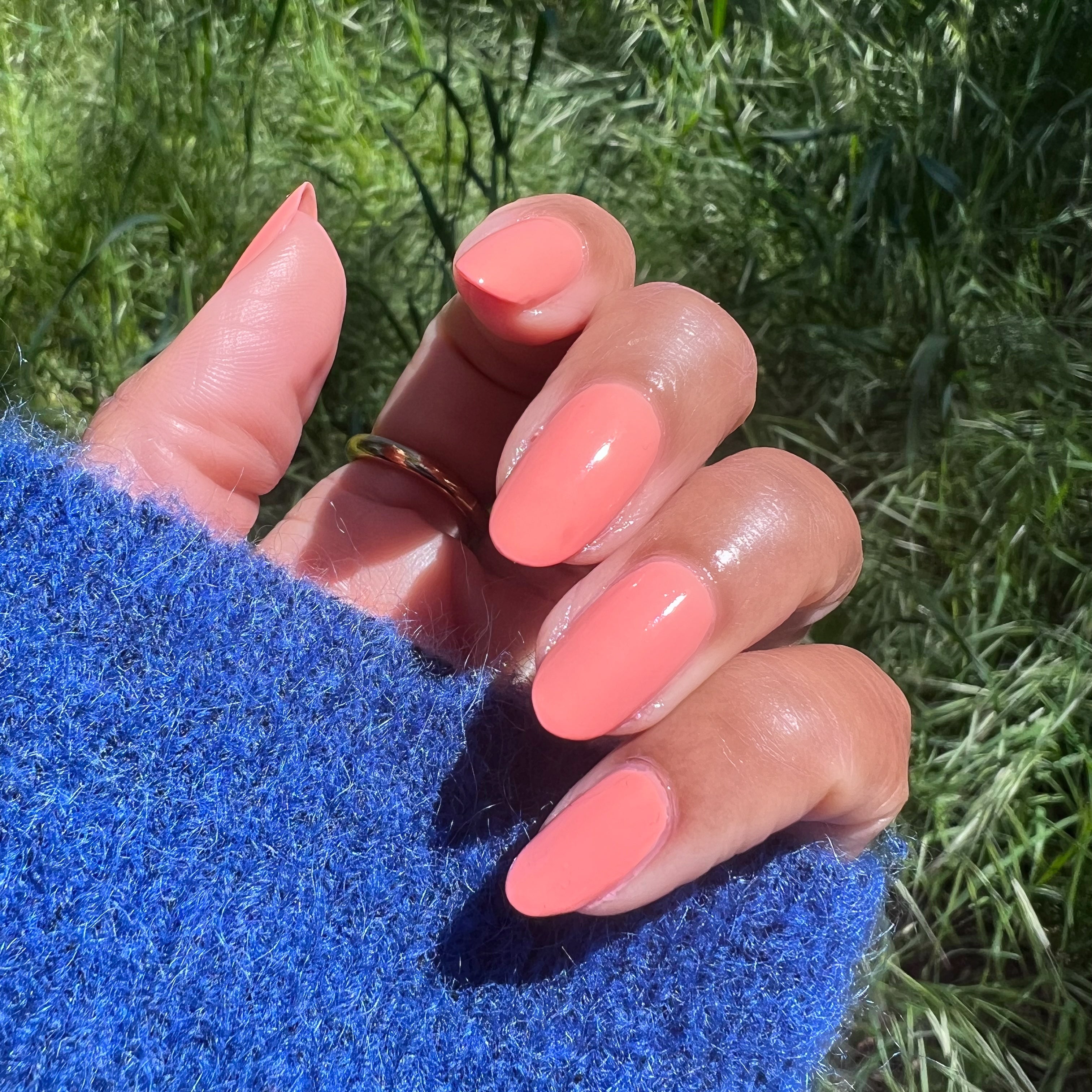 Hand with oval nails painted in glossy Melon quick dry polish outdoors against green grass background