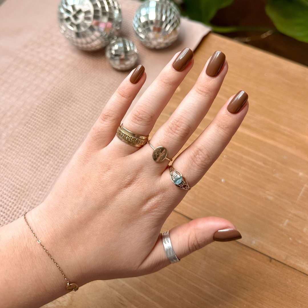 Americano | Short | Squoval - Tab Press-On showing hand with short coffee brown false nails and gold rings on wooden surface