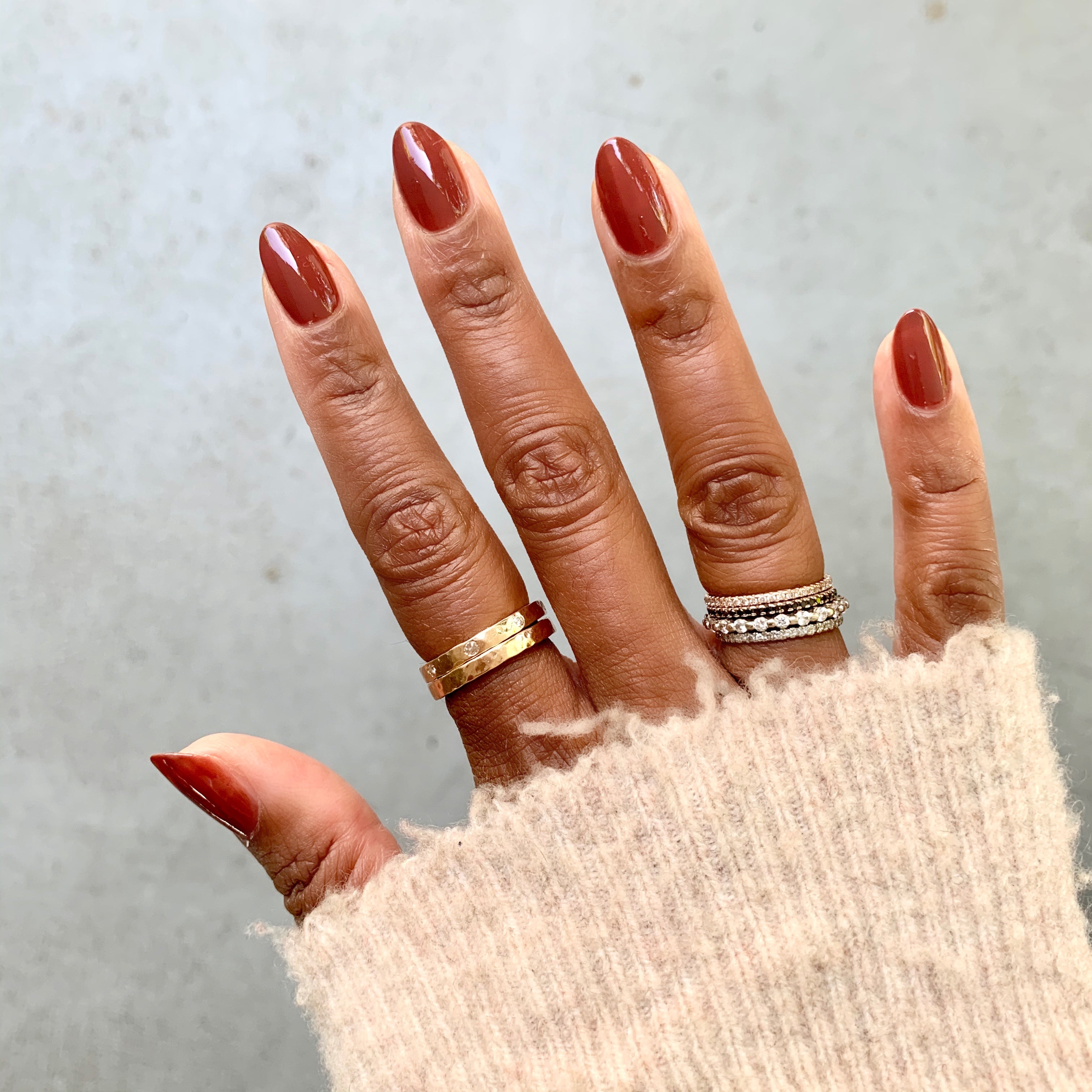 Hand with nails painted in JJ gel polish, a warm cinnamon brown, styled with gold and silver rings and cozy sweater sleeve