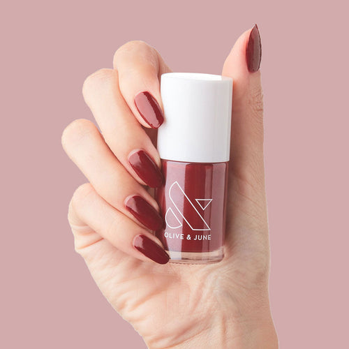 Hand holding a bottle of Cozy Up nail polish with nails painted in warm brick red for a cozy manicure look