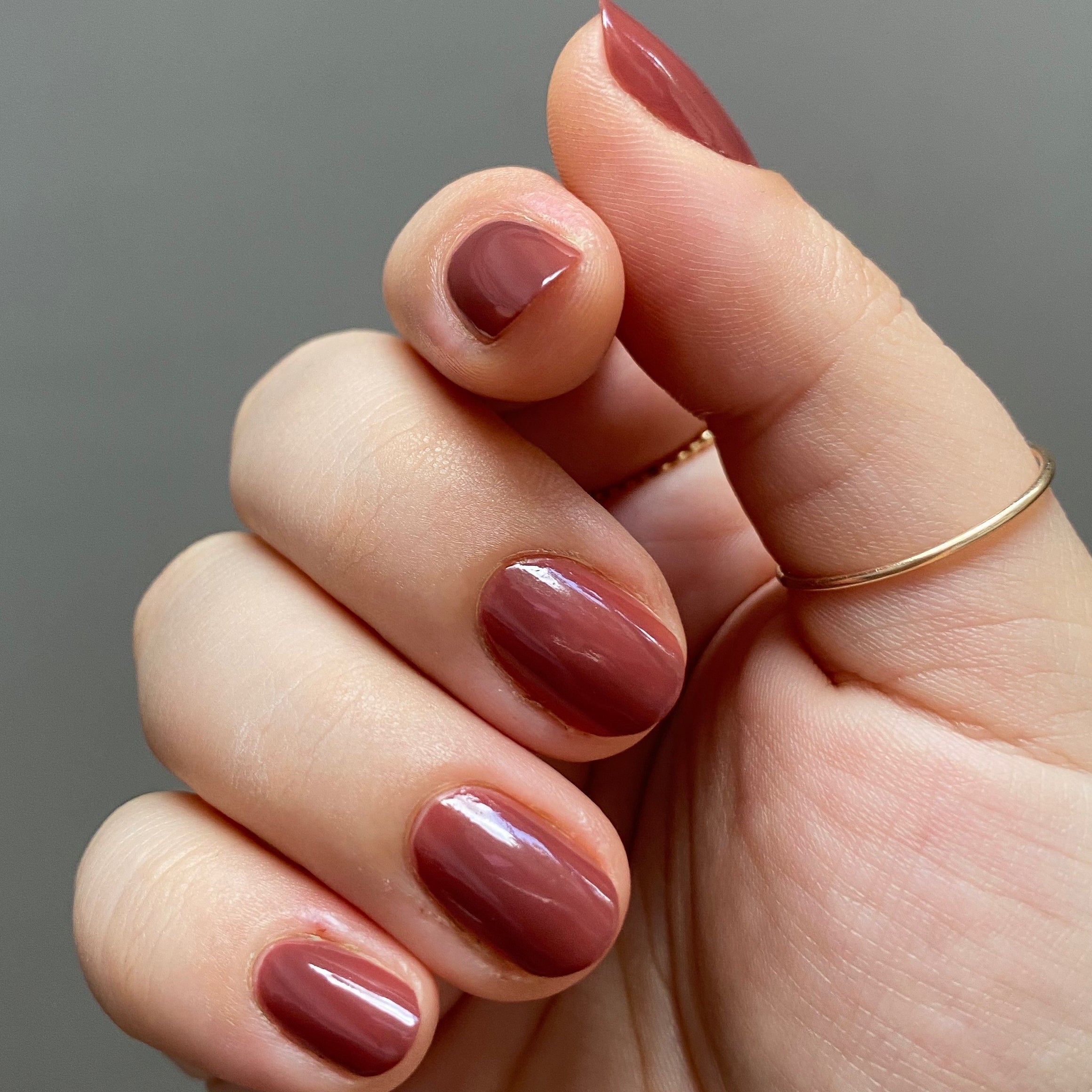 Close-up of hand with nails painted in JJ, a warm cinnamon brown gel polish, wearing a thin gold ring for a simple mani look