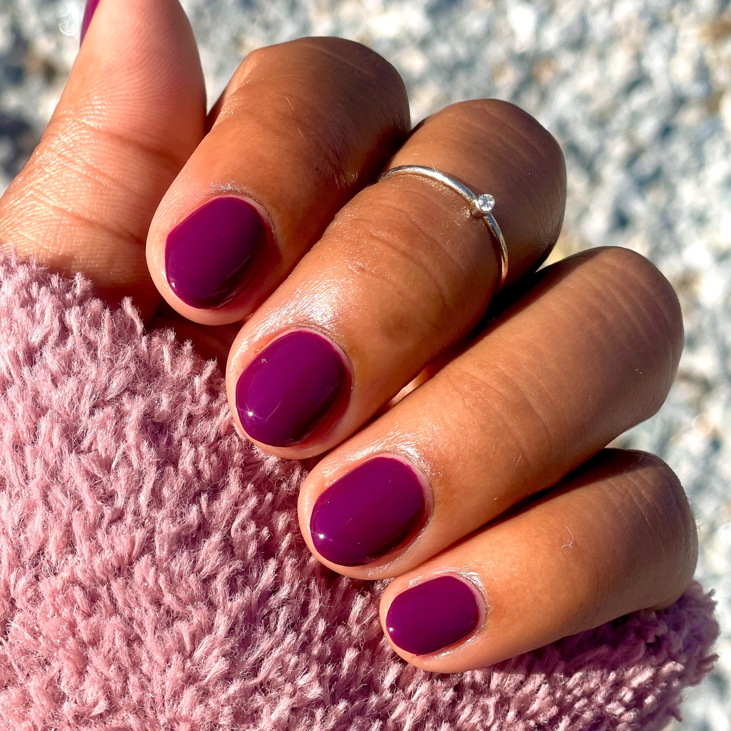 Hand with short nails painted in glossy Plaza deep mulberry purple gel polish against soft textured fabric background