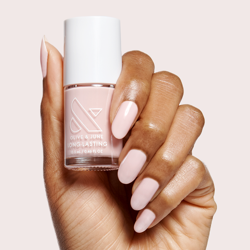 Hand with soft sheer pink manicure holding Dream Job nail polish bottle against pale pink background