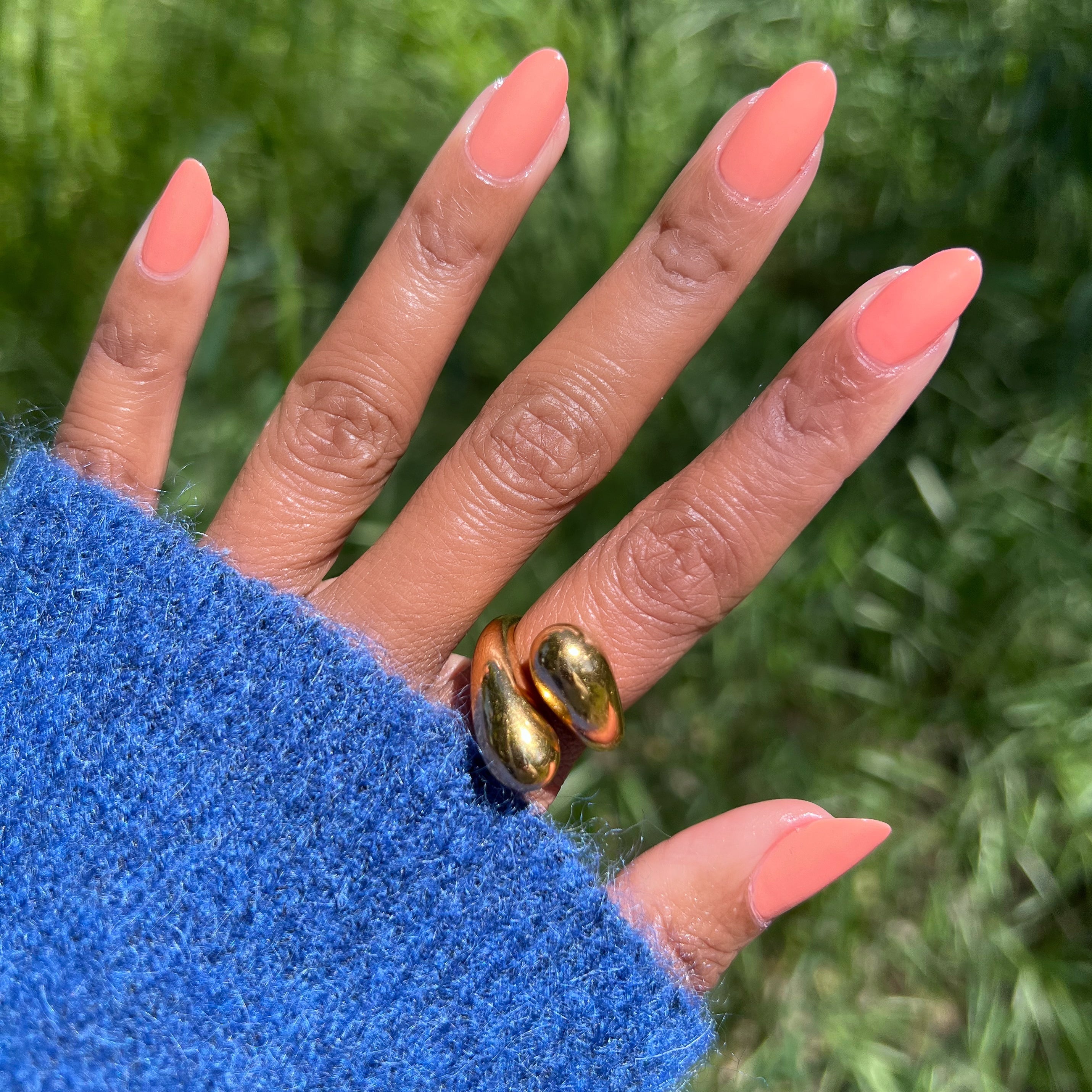 Hand with almond-shaped nails painted in Melon quick dry polish showing gold rings outdoors with green grass background