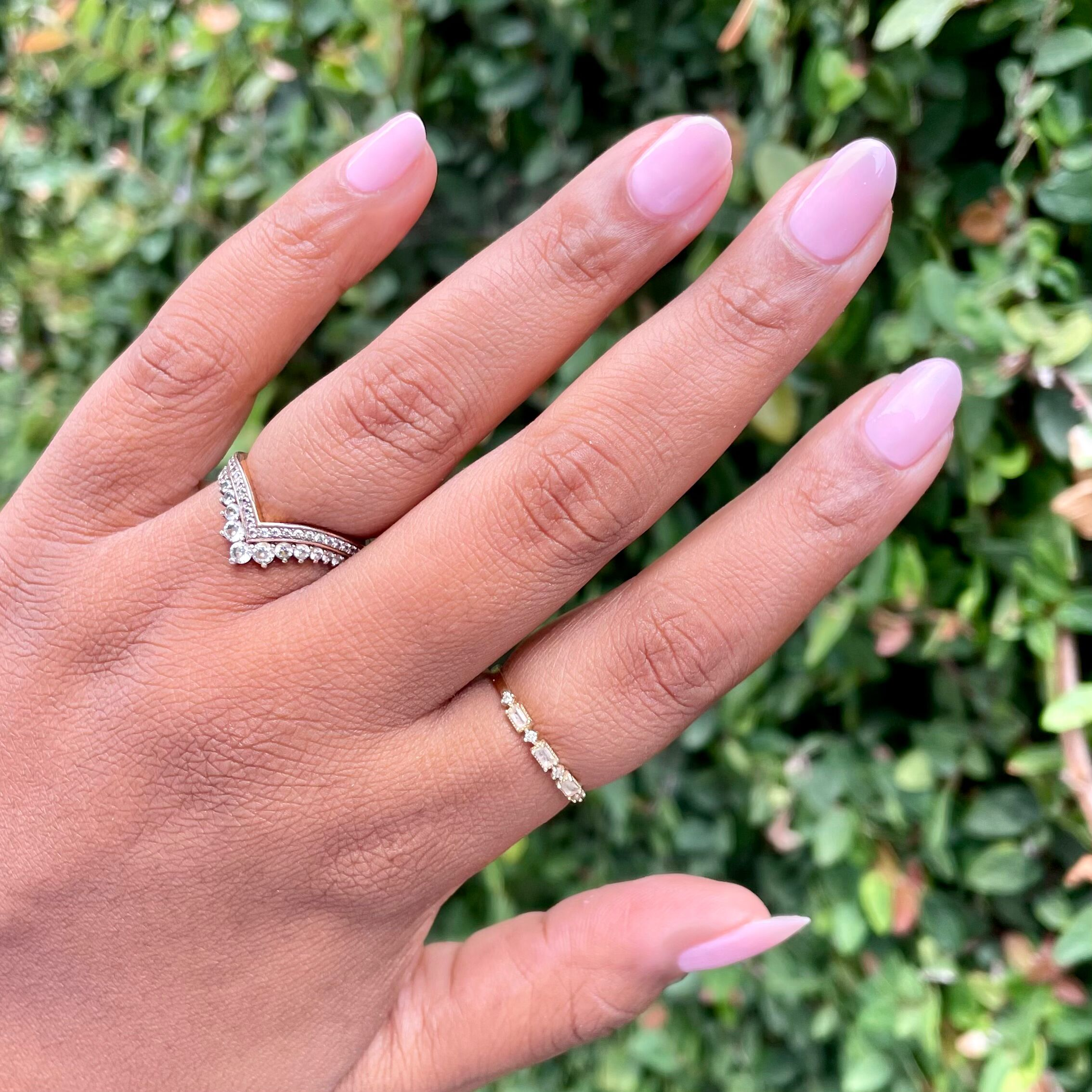 Hand with Dream Job sheer milky pink manicure wearing two delicate rings against a green leafy backdrop