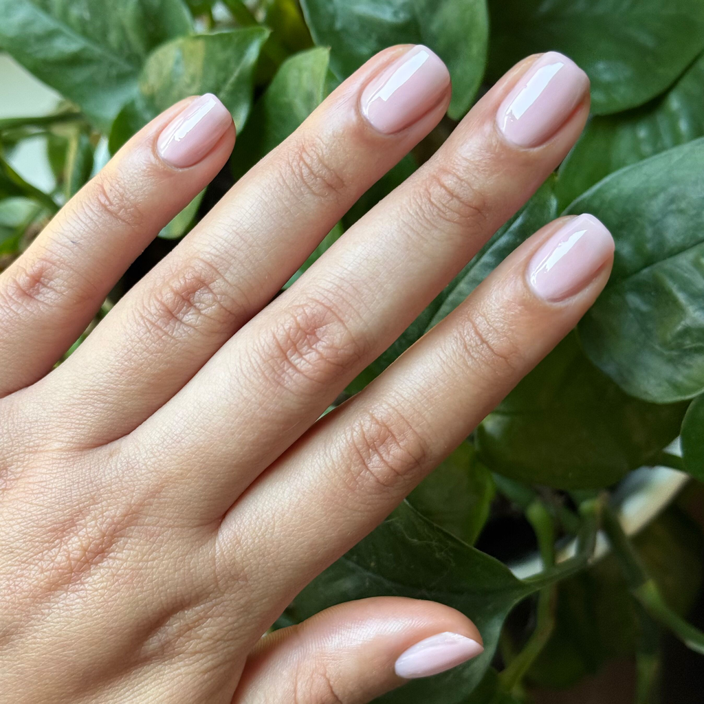 Hand with glossy sheer neutral pink manicure wearing Dream Job polish against green leafy background