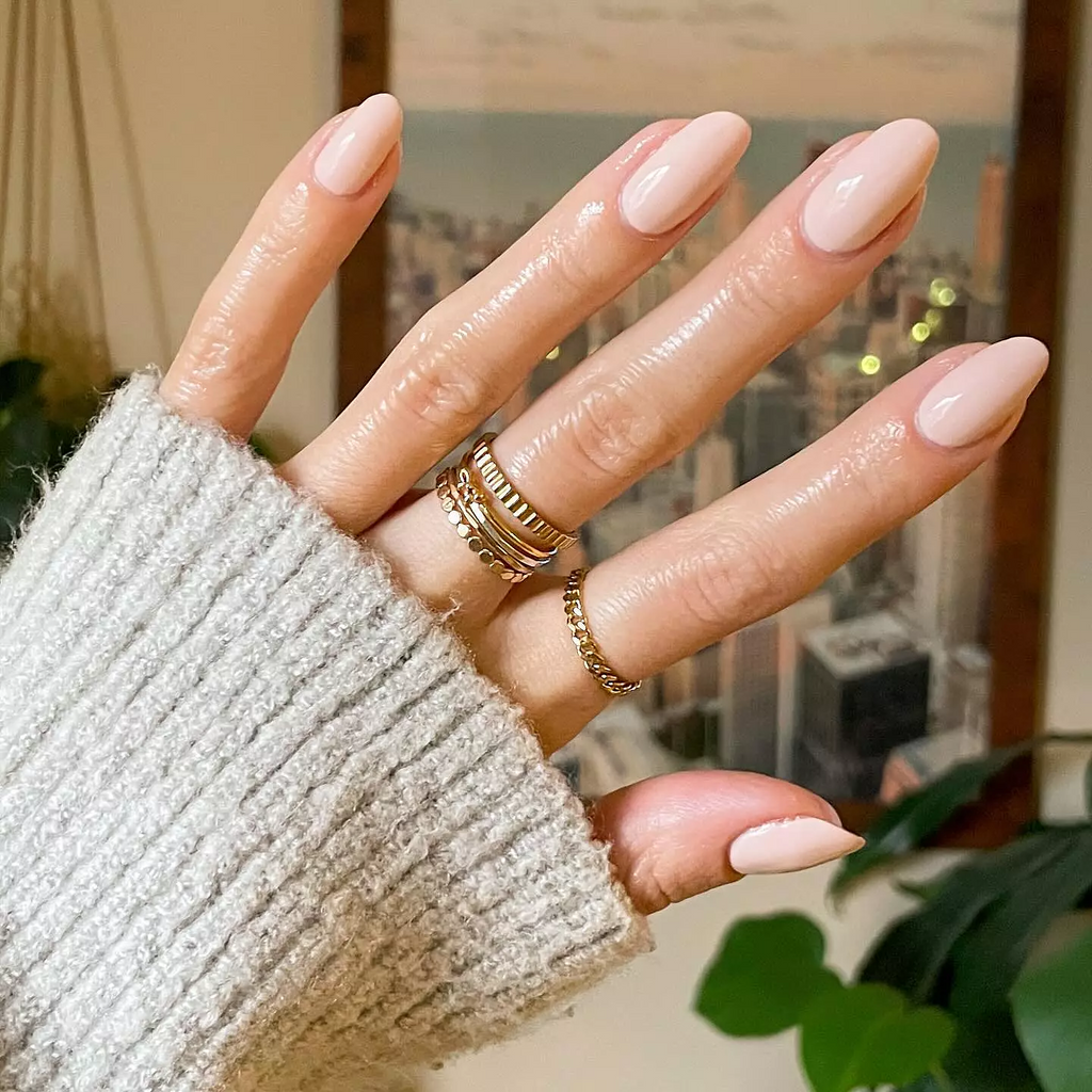 Hand with smooth neutral peachy pink KAG gel polish on nails worn with gold rings against a blurred background