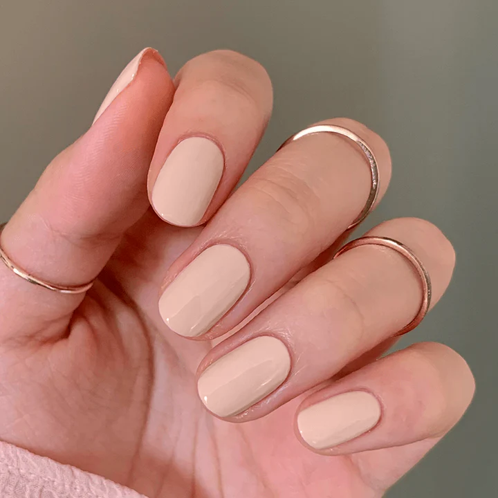 Hand with KAG neutral peachy pink gel polish on nails wearing delicate rose gold rings against a soft neutral background