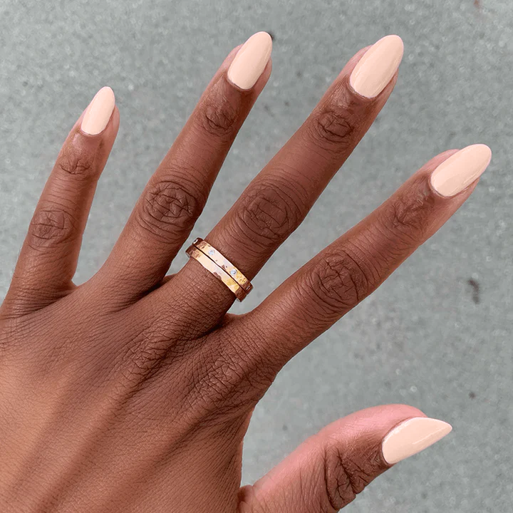 Hand with smooth KAG gel polish in neutral peachy pink on nails wearing a gold ring against a gray background