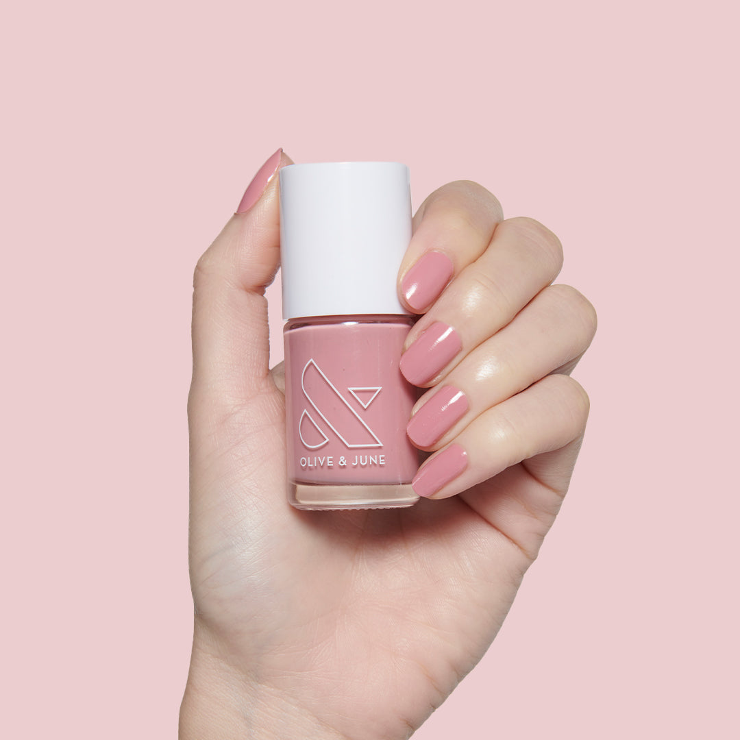 Hand with neatly manicured nails painted in dusty rose pink Yogurt Parfait polish holding matching bottle against pink background