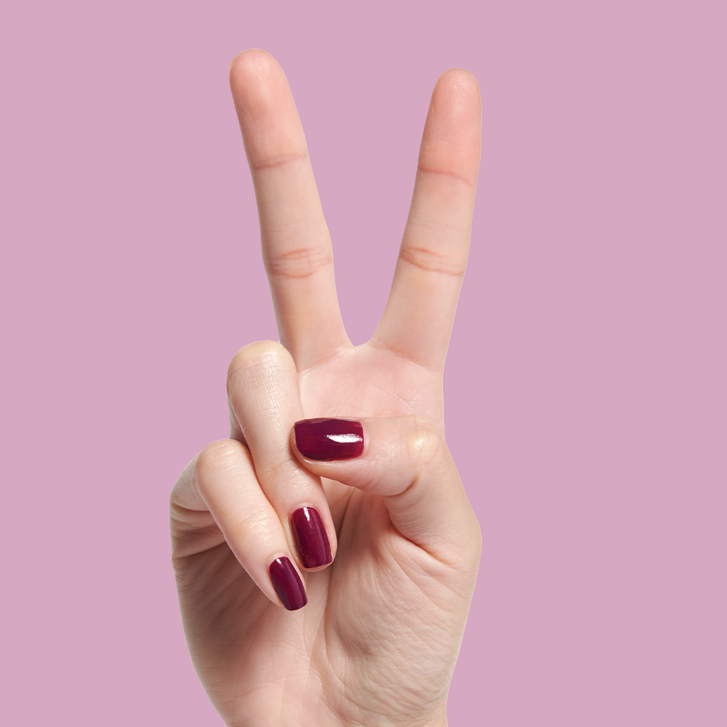 Hand with short nails painted in deep mulberry purple Plaza gel polish against a soft pink background