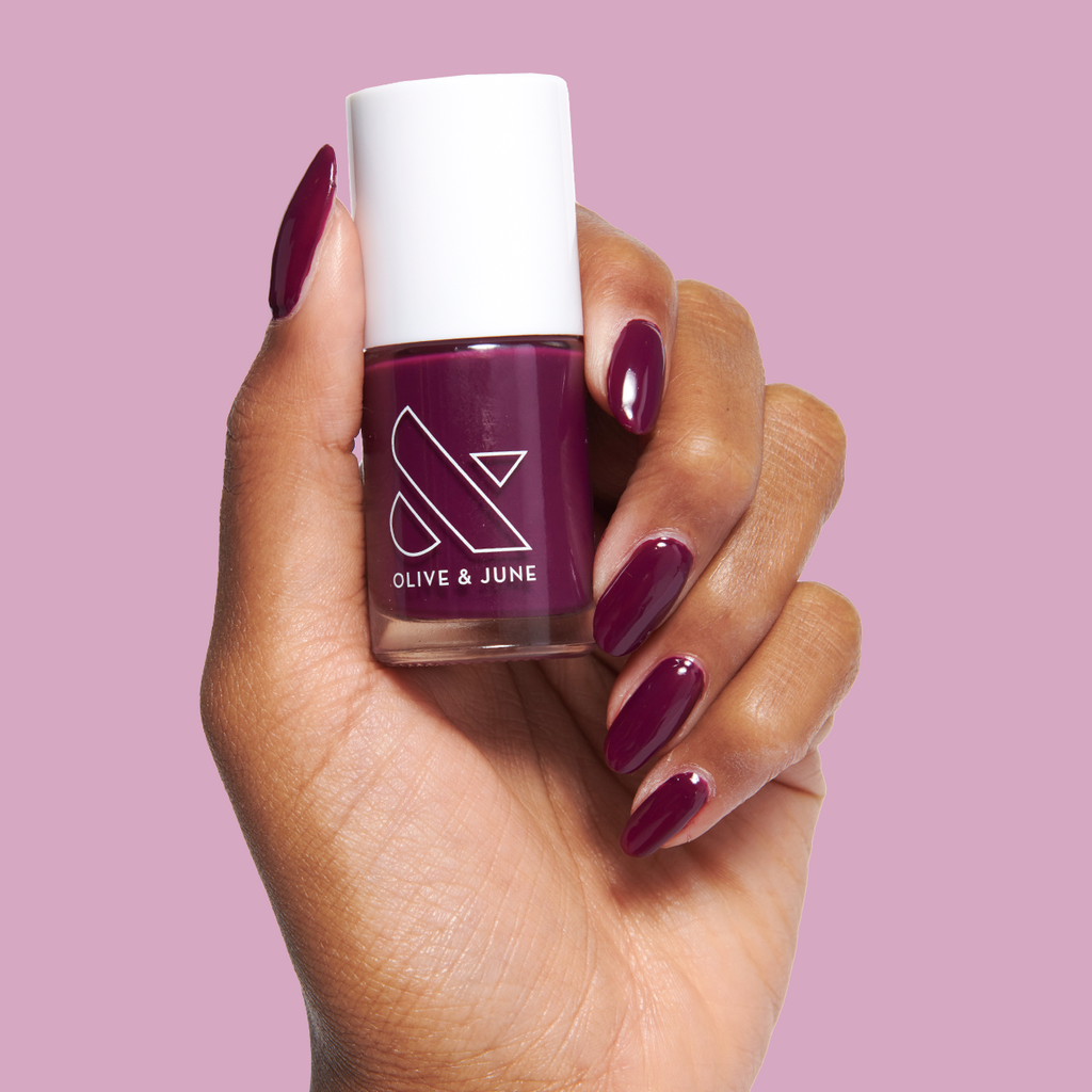Hand holding Plaza nail polish bottle with deep mulberry purple gel polish on glossy manicured nails on pink background