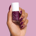 Hand holding Plaza nail polish bottle with deep mulberry purple gel polish on glossy manicured nails on pink background