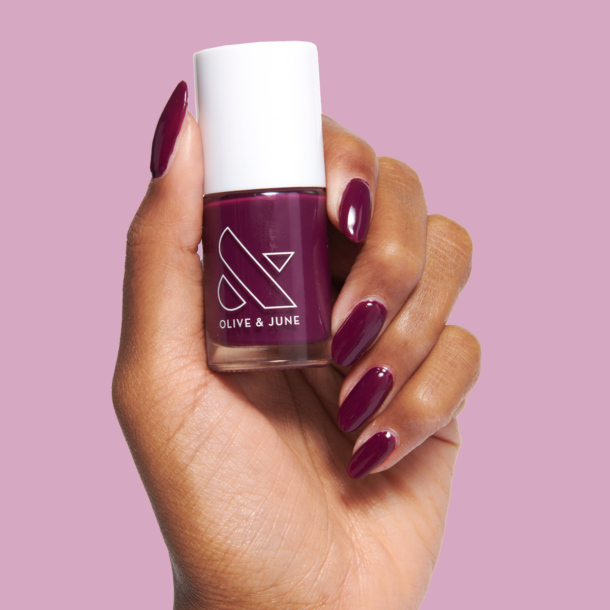 Hand holding Plaza nail polish bottle with deep mulberry purple gel polish on glossy manicured nails on pink background
