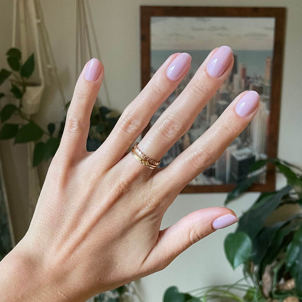 Hand with short rounded nails painted in Passionfruit Gloss cool milky lavender gel polish indoors with plants and city artwork background