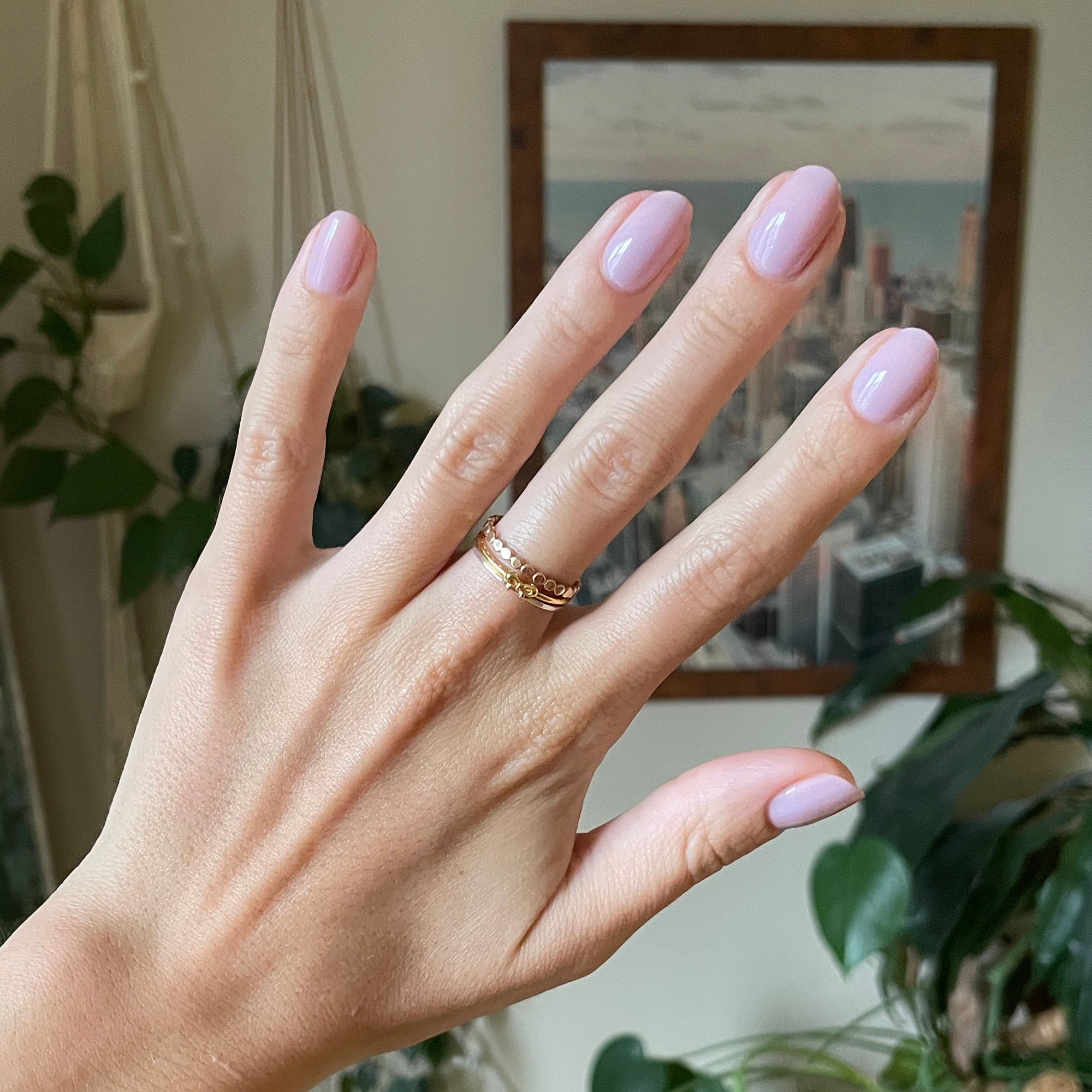 Hand with short rounded nails painted in Passionfruit Gloss cool milky lavender gel polish indoors with plants and city artwork background