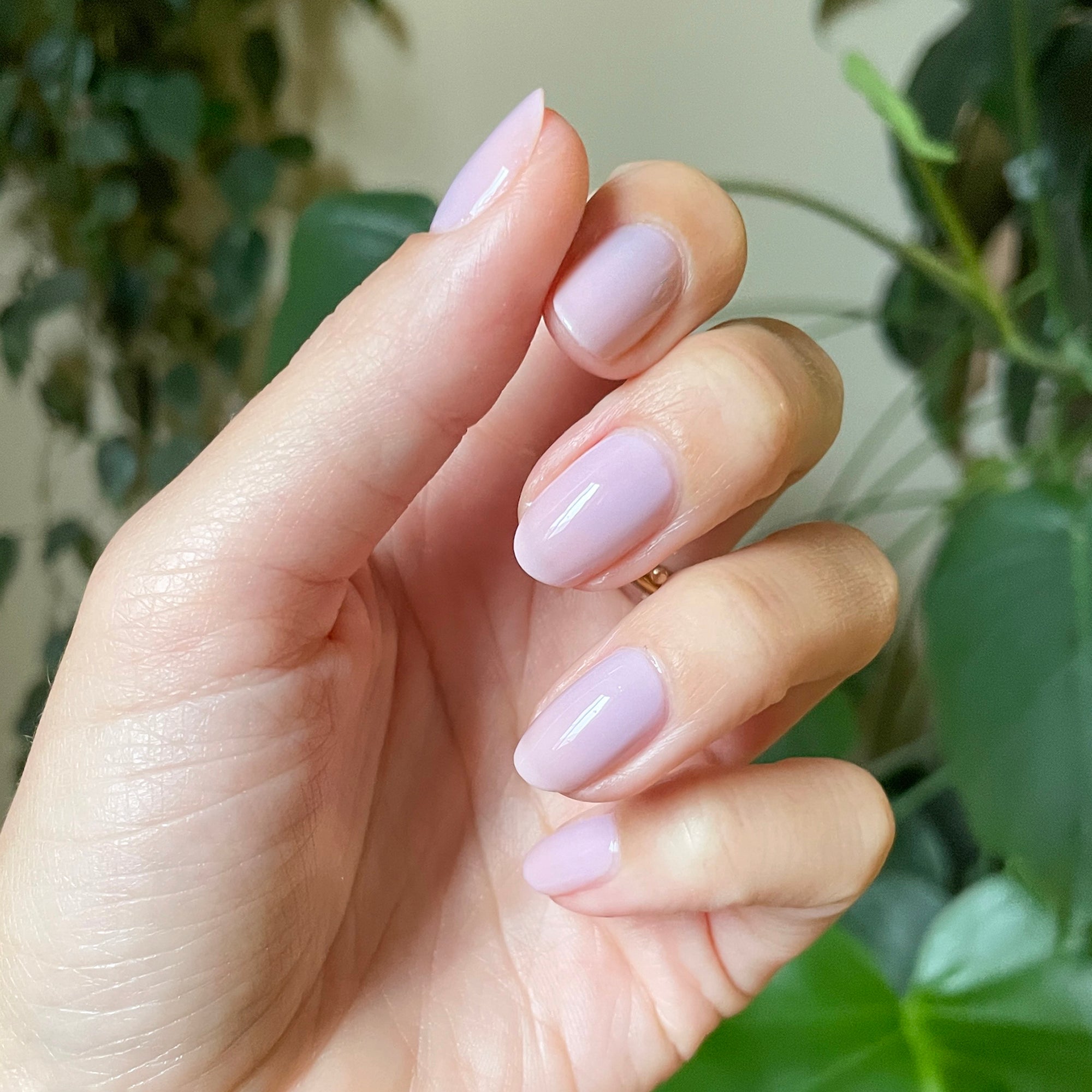 Hand with short almond-shaped nails painted in glossy Passionfruit Gloss milky lavender gel polish indoors with green plants background
