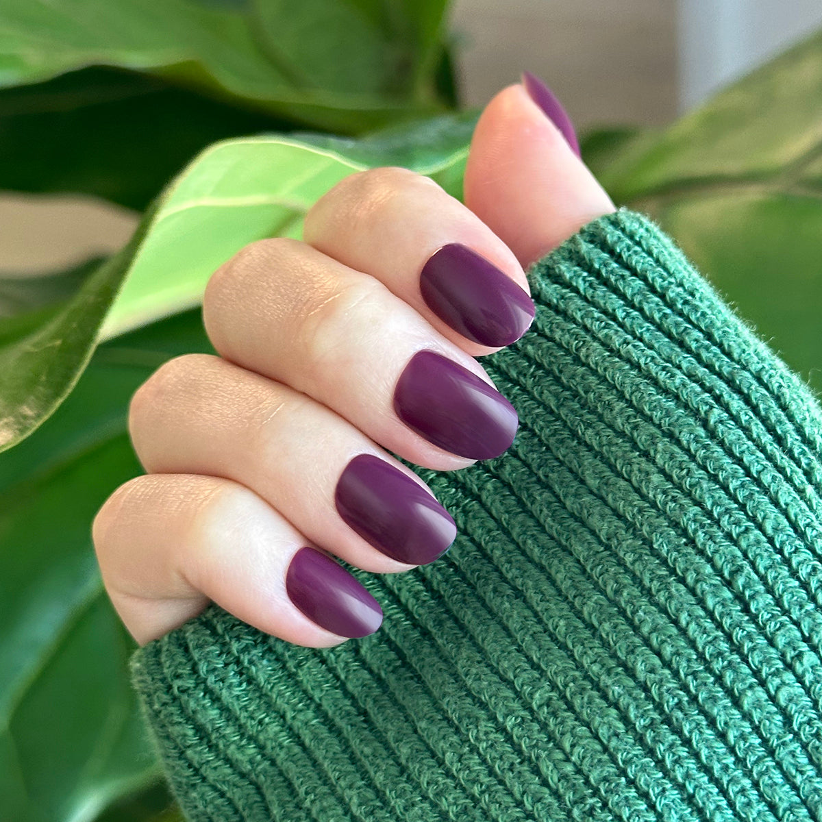 Hand with extra short round nails in dark purple shade Who Likes Raisins? - Press-On | Extra Short | Round worn with green sweater sleeve