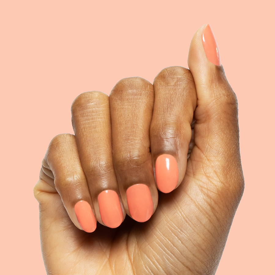 Hand with medium nails painted in bright coral Ride into the Sunset gel polish against a soft peach background