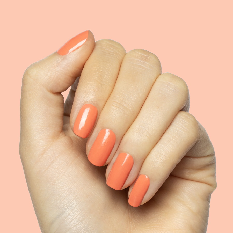 Hand with nails painted in coral Ride into the Sunset gel polish on medium-length nails against peach background