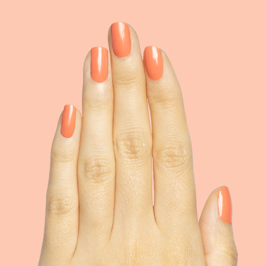 Hand with nails painted in coral Ride into the Sunset gel polish on medium-length nails against soft peach background