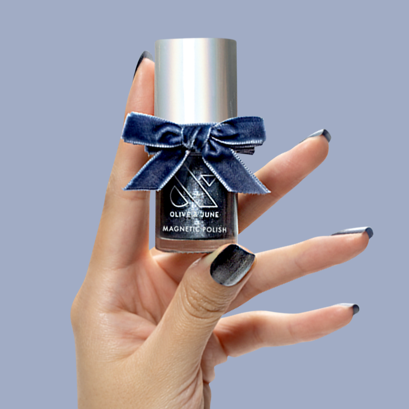 Hand holding Olive and June Sapphire Velvet magnetic polish bottle with icy blue nails on lavender background