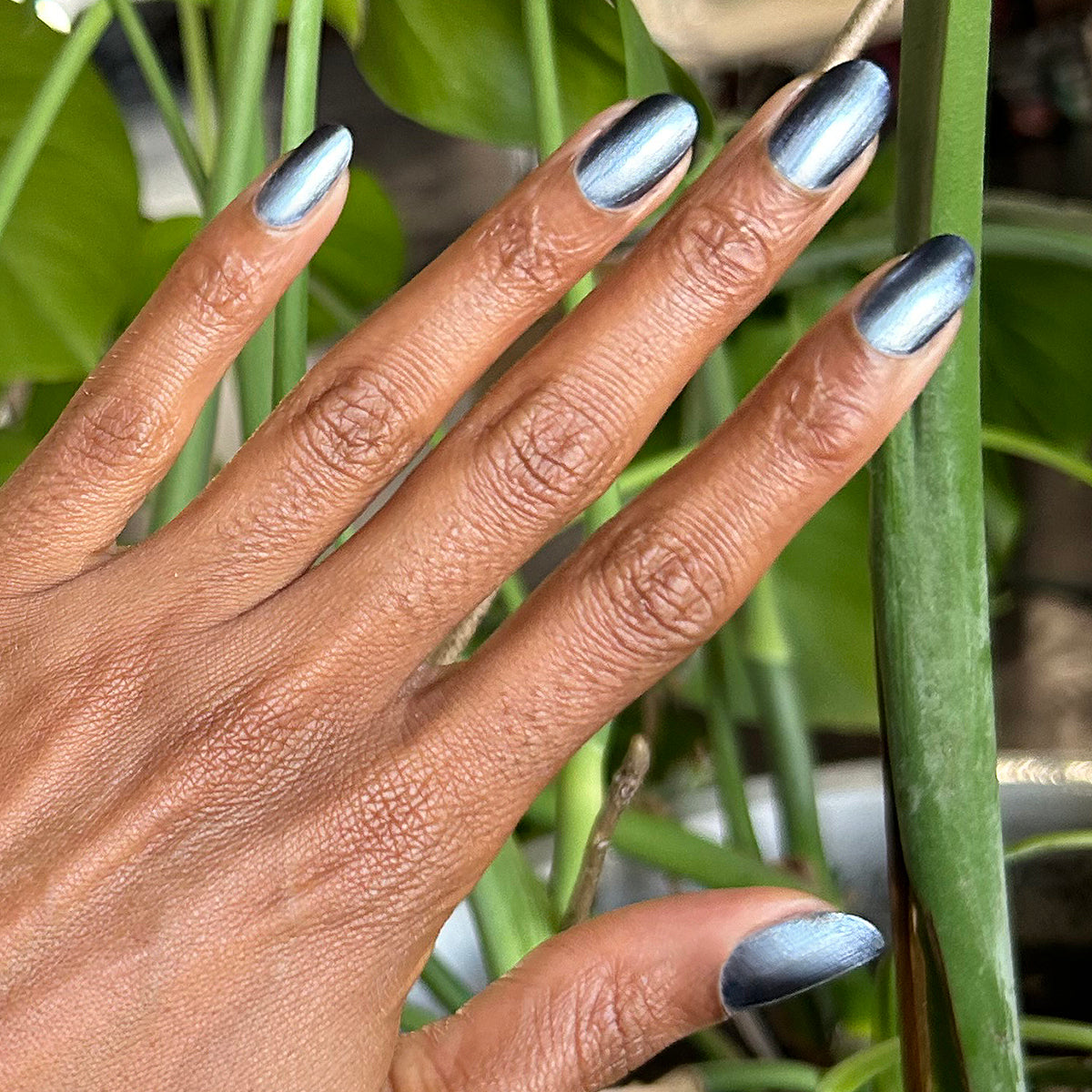 Hand with nails painted in Sapphire Velvet icy blue magnetic polish showing gradient sheen for manicure with plants background
