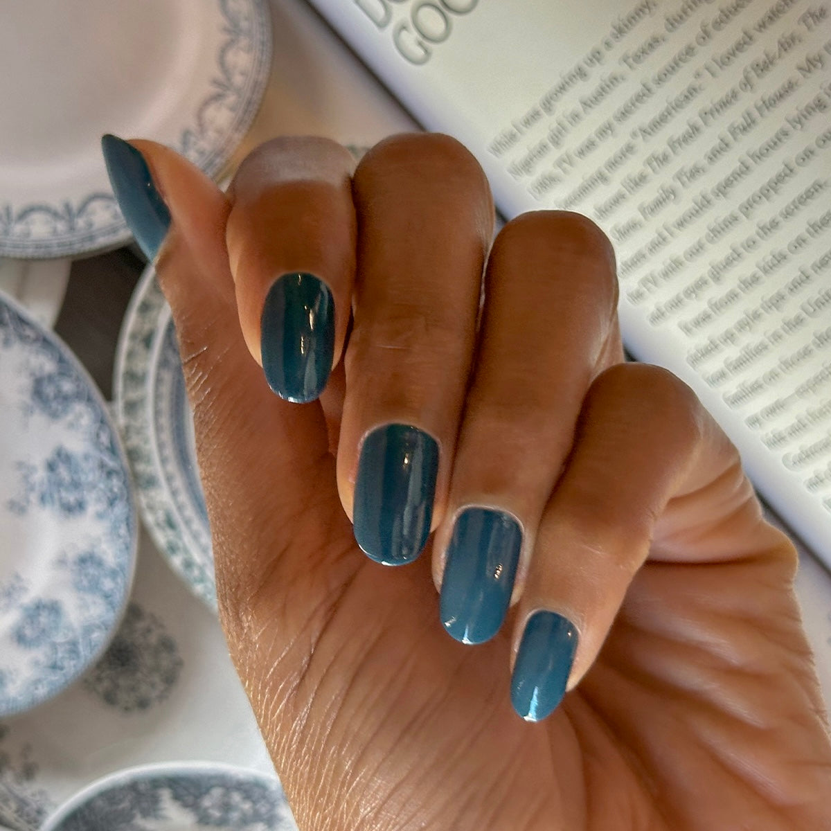 Hand with Bodysuit gel polish manicure in deep dusty cool teal on medium-length nails over vintage blue and white plates background