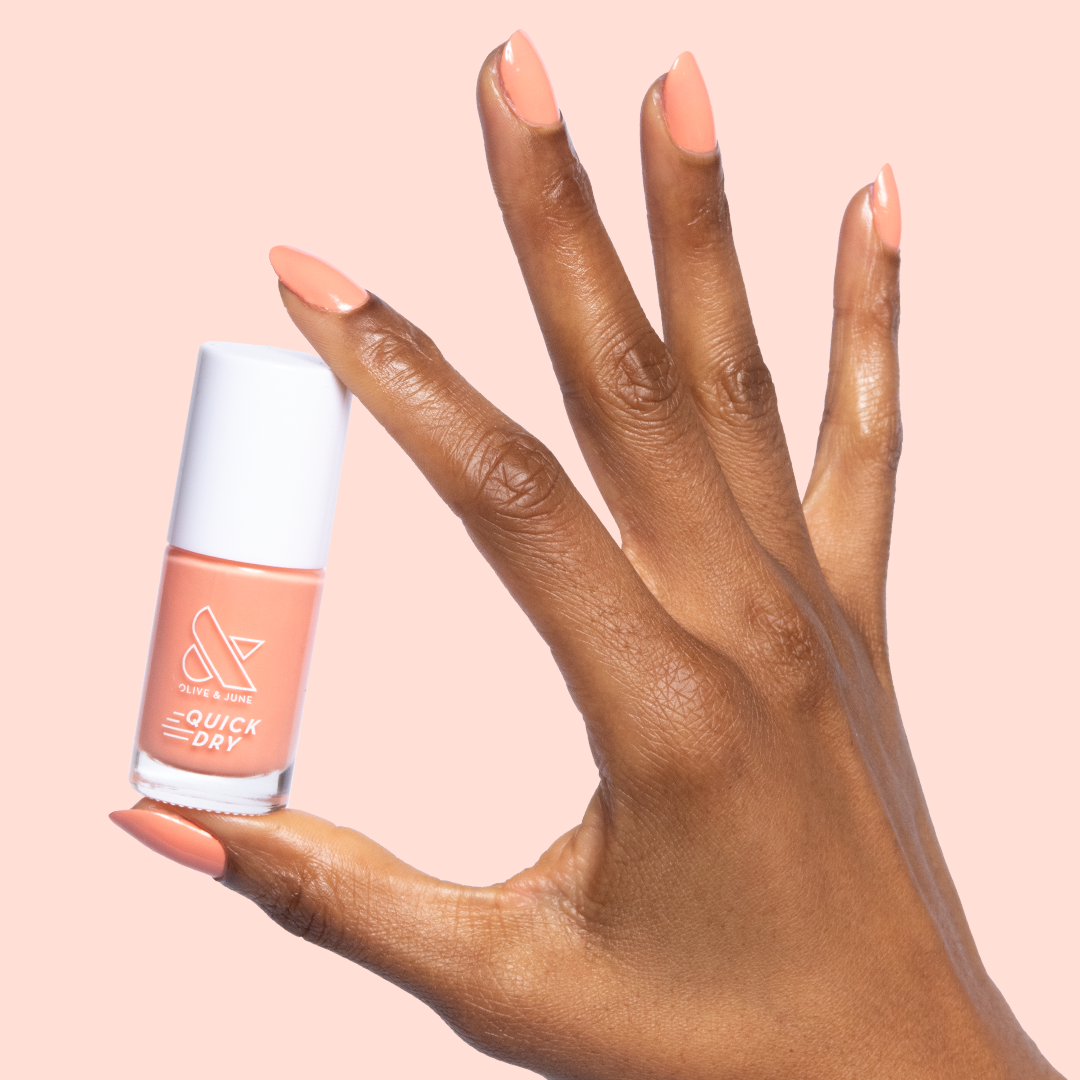 Hand with nails painted in peachy Melon quick dry polish holding matching bottle on a pale pink background