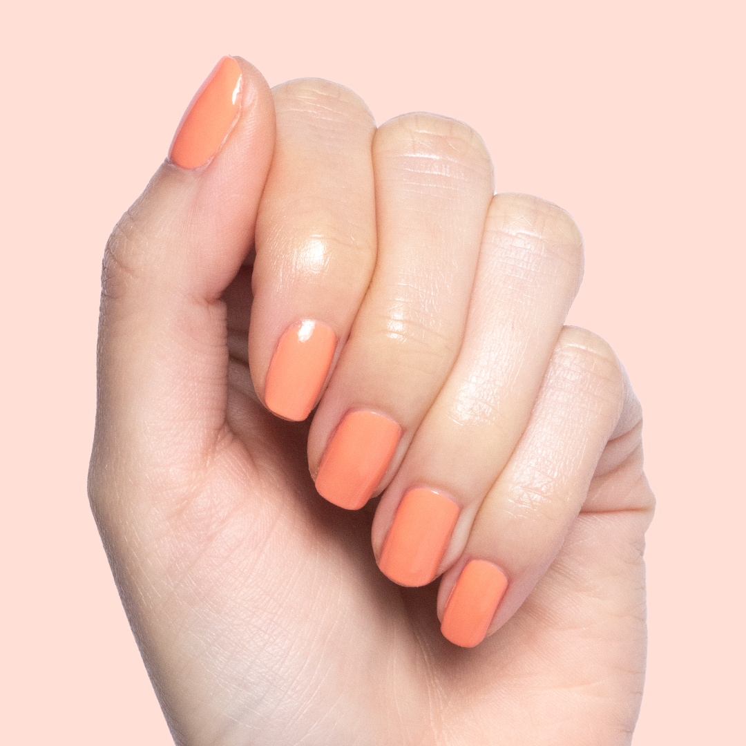 Hand with short nails painted in glossy Melon quick dry polish against a soft peach background