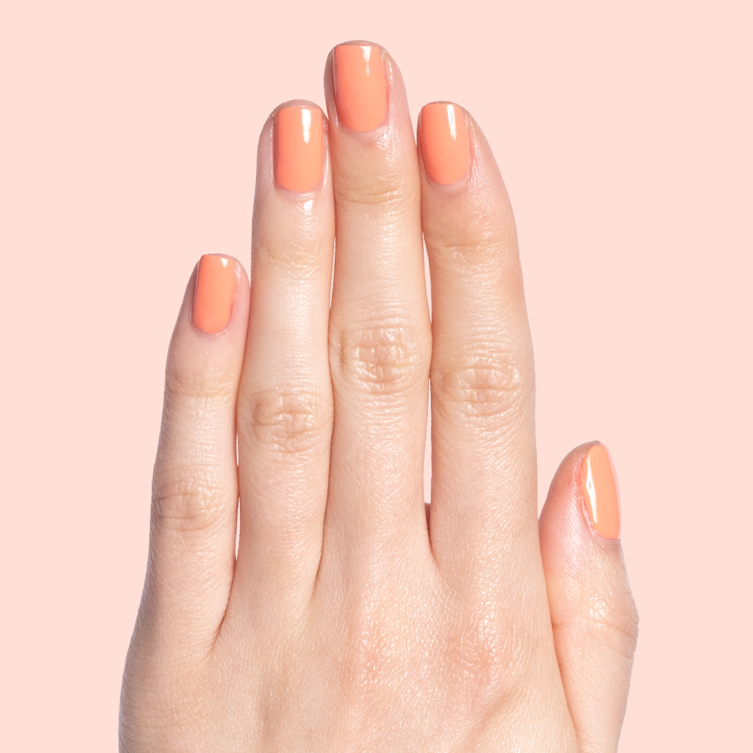 Hand with nails painted in glossy Melon quick dry polish in a soft cantaloupe shade on light peach background