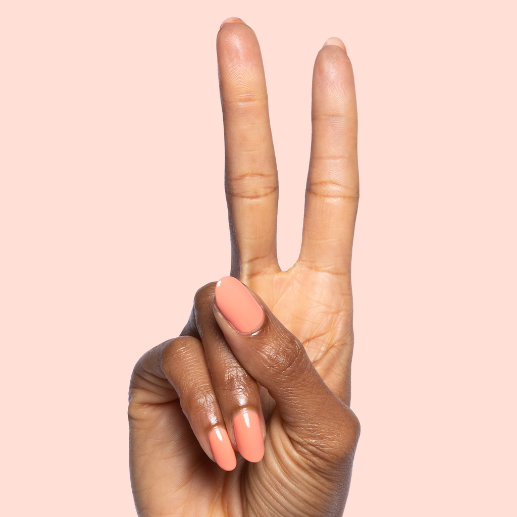 Hand with almond-shaped nails painted in glossy Melon quick dry polish showing peace sign on pale peach background