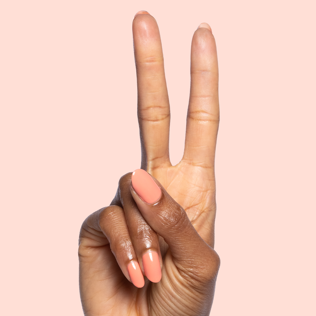 Hand with almond-shaped nails painted in glossy Melon quick dry polish showing peace sign on pale peach background