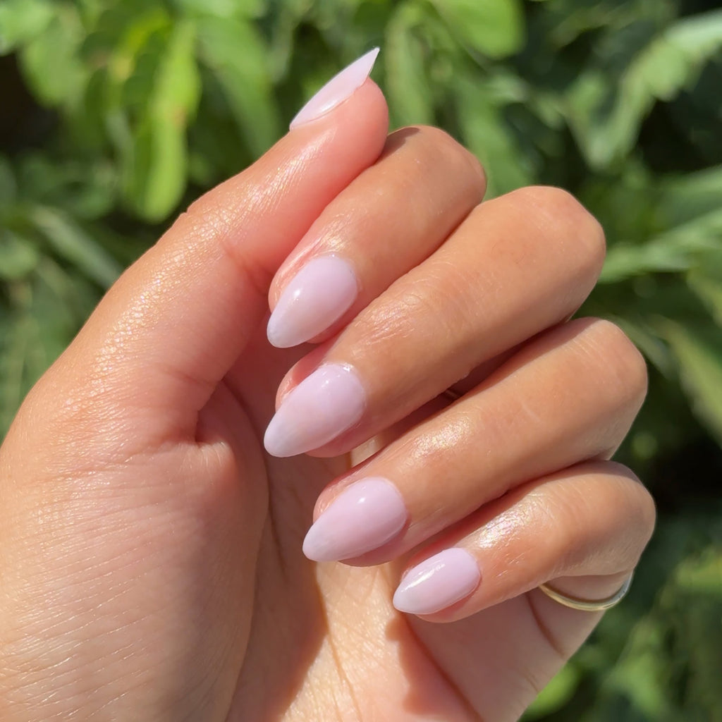 Hand with almond-shaped nails painted in milky lavender using Passionfruit Gloss gel polish outdoors with leafy green background
