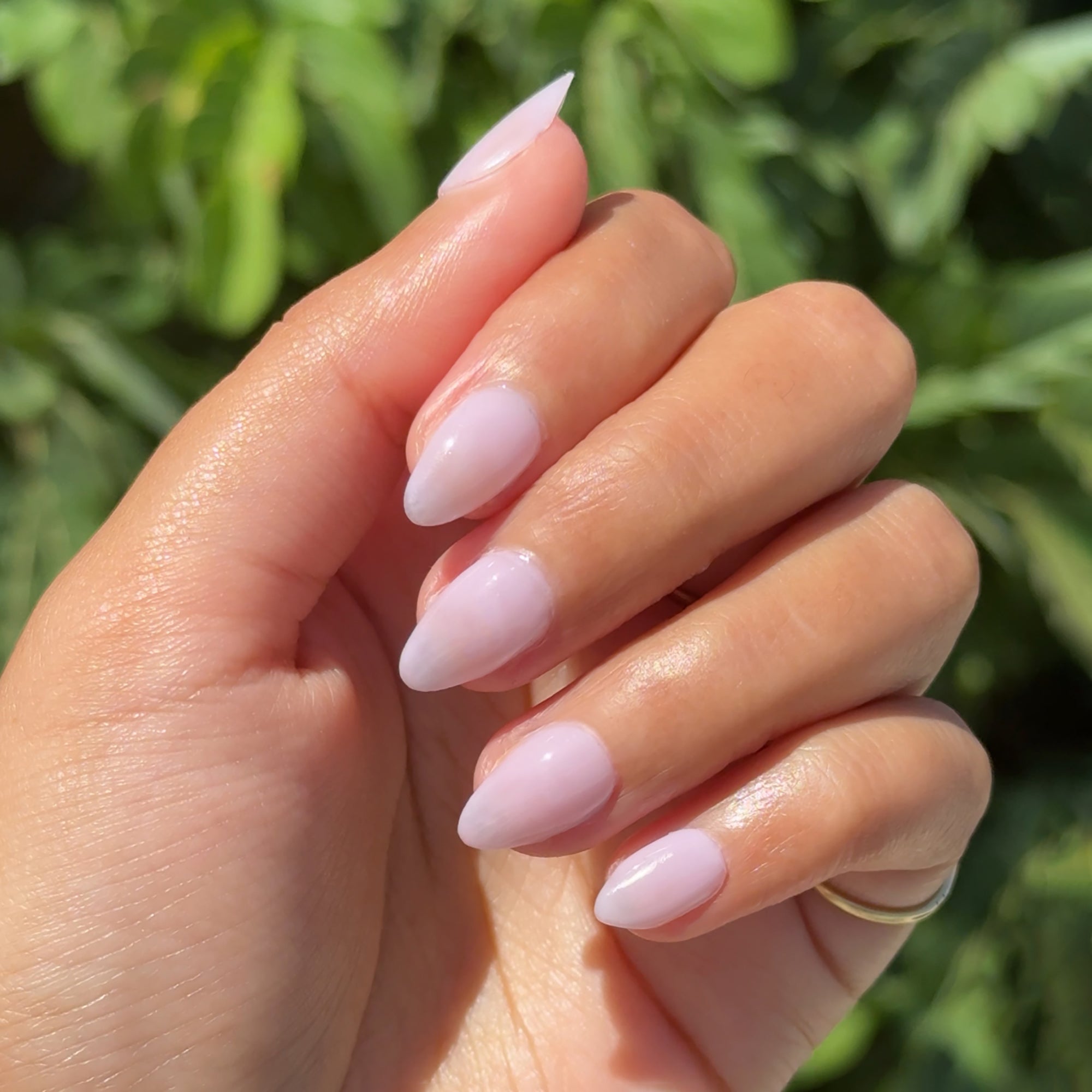 Hand with almond-shaped nails painted in milky lavender using Passionfruit Gloss gel polish outdoors with leafy green background
