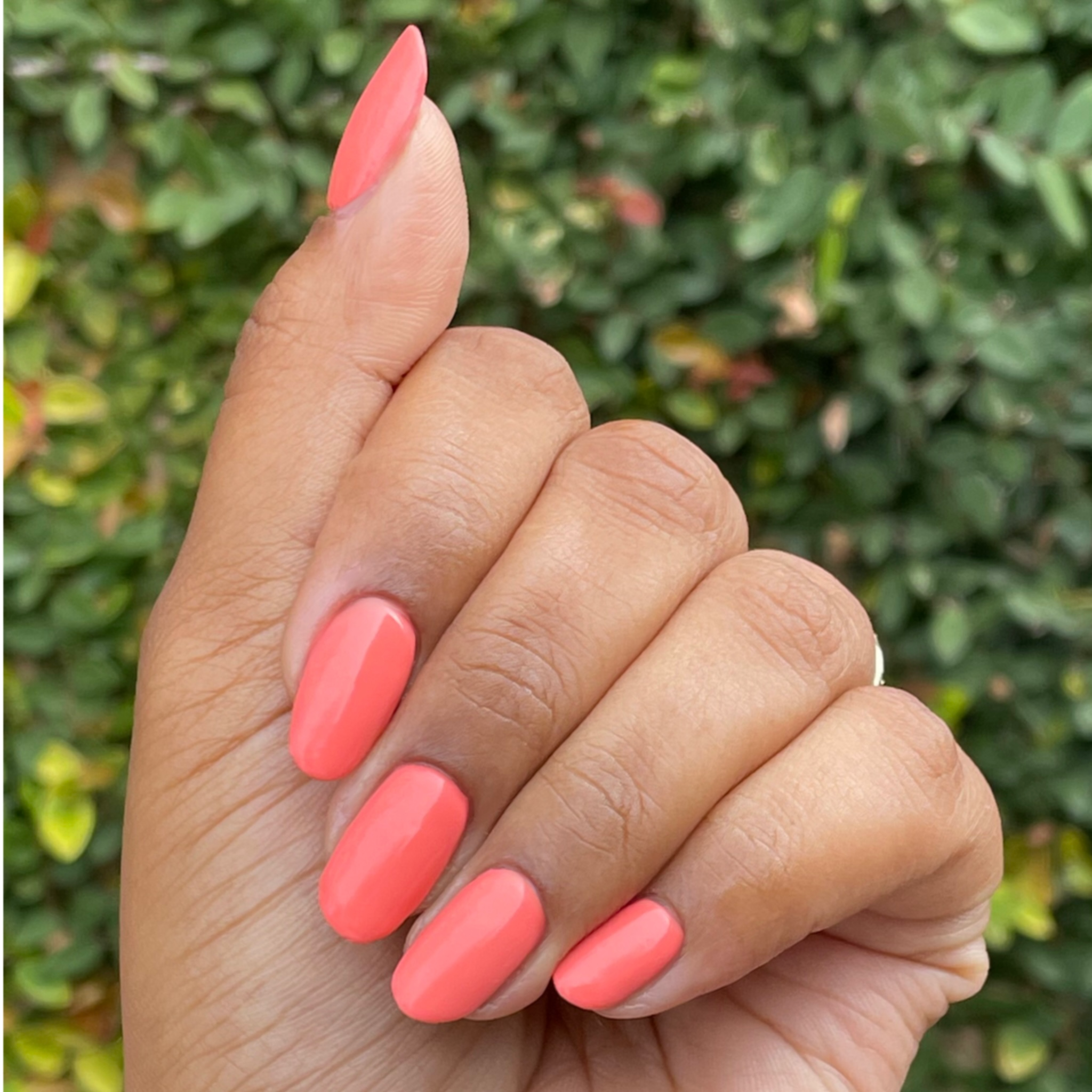 Hand with almond-shaped nails painted bright coral Ride into the Sunset gel polish outdoors with blurred greenery background
