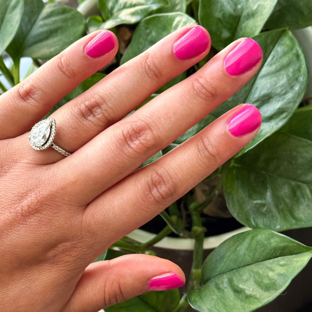 Hand with nails painted vibrant magenta using Wild Orchid - Gel for a bold, long-lasting mani against green plant background